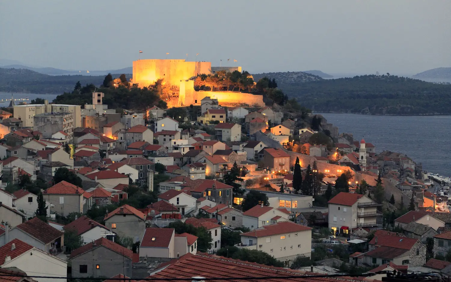 The old town of Sibenik at dusk with the floodlit fortress of St Michael above the red roofs and the channel beyond, Croatia